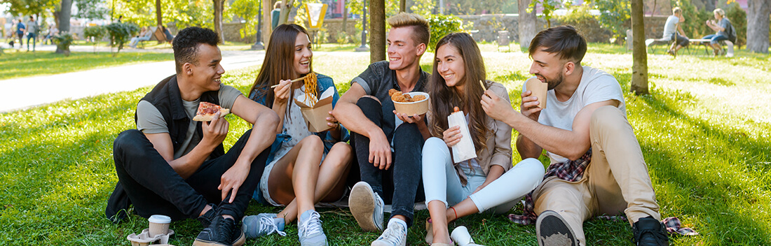A group of young people smiling, sitting outside and eating a variety of foods