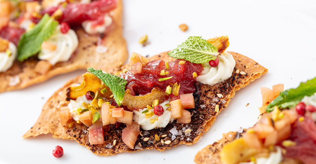 Crispy cracker topped with whipped cream cheese, diced apples, mint, and a berry sauce with nuts and seeds